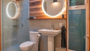 A modern bathroom with a glass shower enclosure, a white toilet, a pedestal sink, and a round mirror with lights.