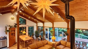 A cozy, well-lit wooden interior with a dining area, sofas, and a large window overlooking greenery.