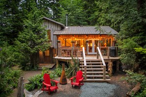 Una cabaña rústica con una terraza de madera, una escalera y dos sillas Adirondack rojas.
