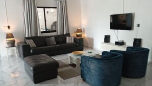 Een moderne woonkamer met een bank, poef en blauwe fauteuils. Er is een flatscreen televisie aan de muur, een salontafel en een lamp.