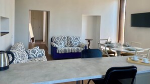 A modern living room with a sofa, a dining table set for two, a TV mounted on the wall, and a large window providing natural light. - Santa Cristina Suites Balcony with a View (Bolsena)