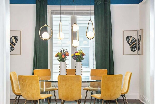 A dining area with a glass table surrounded by four yellow chairs, two vases with flowers, and pendant lights.