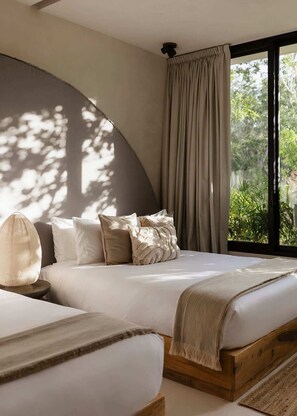 A bedroom with a curved wall, a bed with white bedding, a bedside lamp, and a view of greenery through a window.