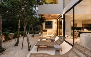 A modern outdoor lounge area with wicker furniture and a glass-enclosed kitchen visible through the glass doors. - Casa Cascada Jungle Villa Beach Club Access (Tulum)