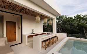 Una cocina exterior moderna con piscina, taburetes de bar y luces colgantes.