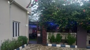 A paved courtyard with a building on one side, a tree with pink flowers, and a blue and white striped barrier. - Bromford Hotel (Owerri)