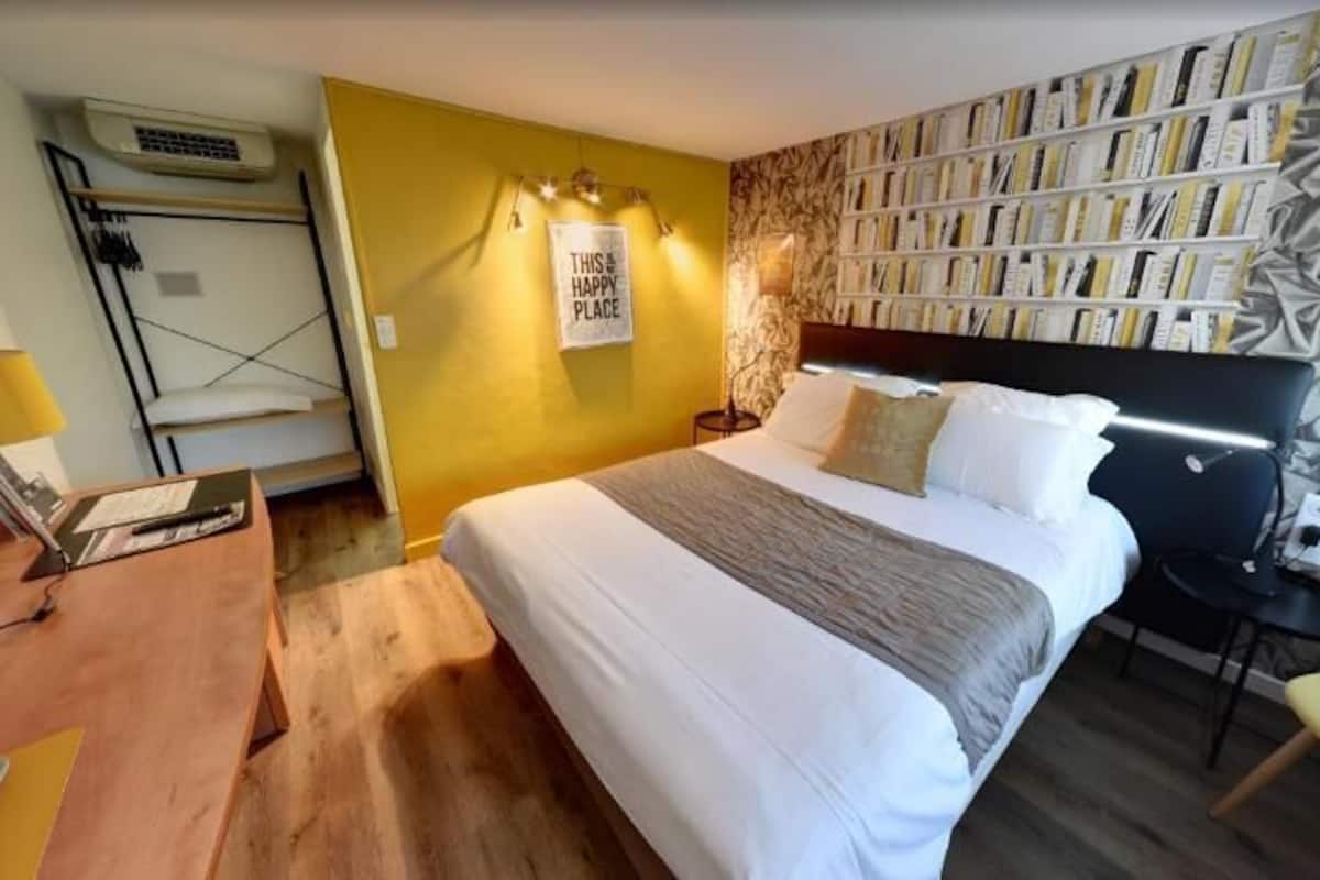 A hotel room with a bed, bedside table, lamp, and a bookshelf.