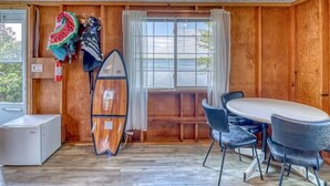 A room with a wooden interior, a table with chairs, a surfboard leaning against the wall, and a window with curtains.