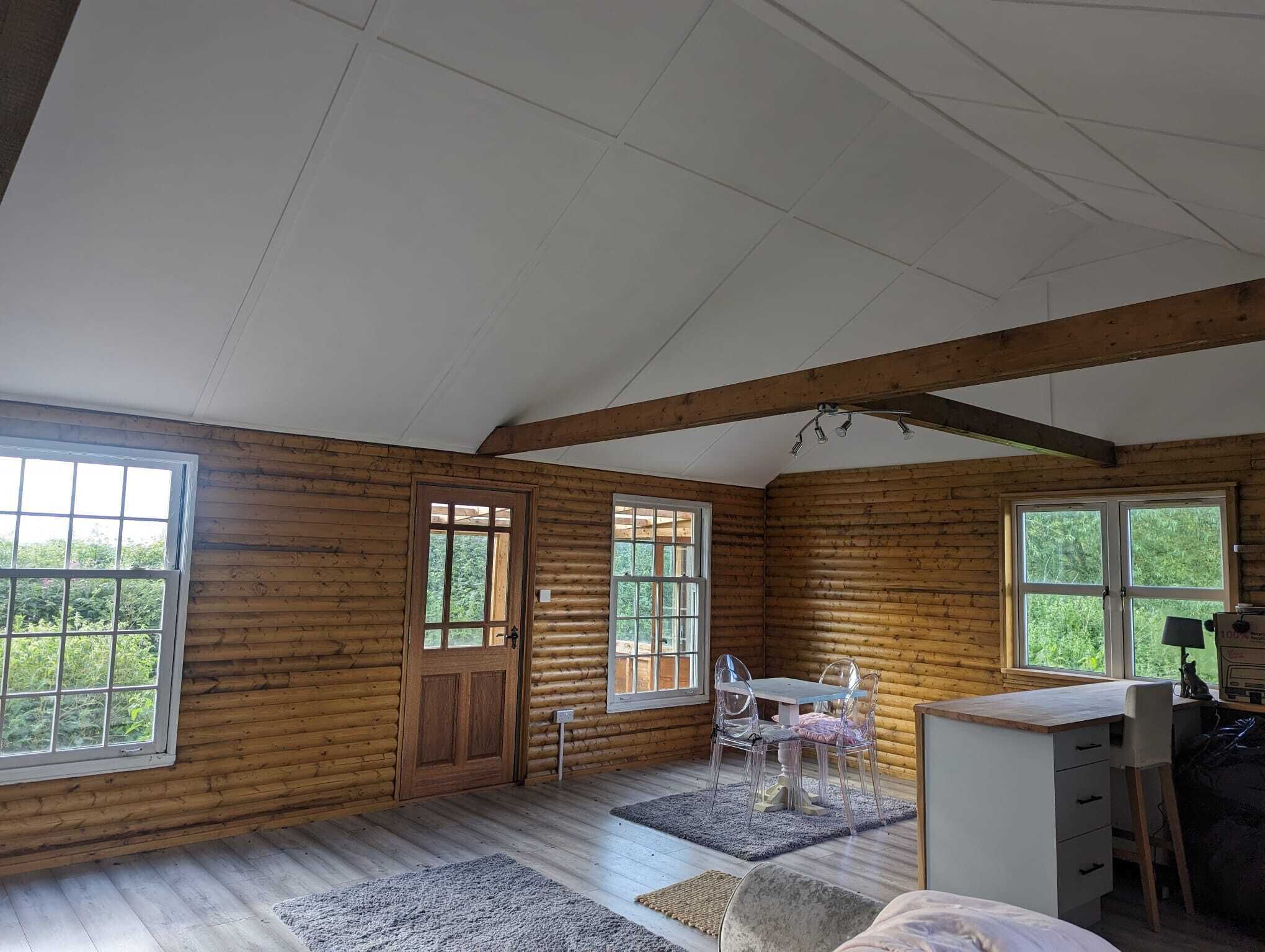 L’intérieur d’une cabane en rondins, avec un plancher en bois, un coin repas comprenant une table et des chaises, un bureau avec une lampe et de grandes fenêtres donnant sur un paysage verdoyant.