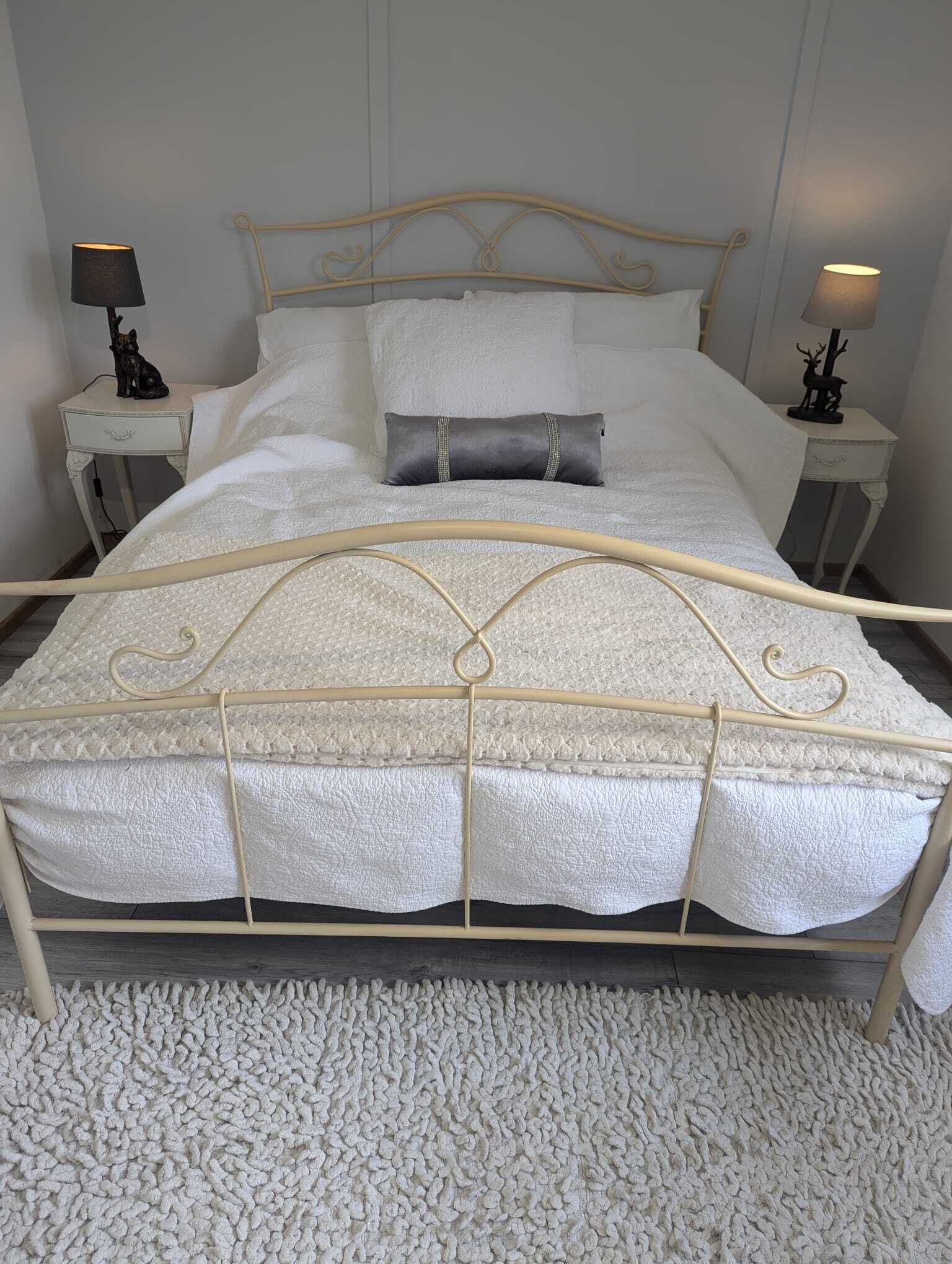 A bed with a white mattress and a beige metal frame, flanked by two bedside tables with lamps and decorative items.