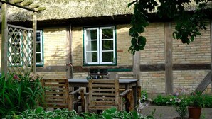 A rustic outdoor dining area with a wooden table and chairs, surrounded by potted plants and a thatched-roof structure. - Judie's garden in Schaalby (Schaalby)