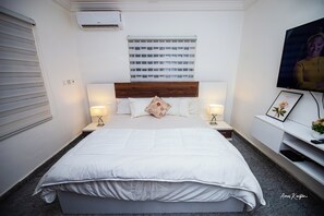 Een keurig opgemaakt slaapkamer met een groot bed, twee nachtkastjes met lampen, een wandgemonteerde televisie en een klein plankje met decoratieve objecten.