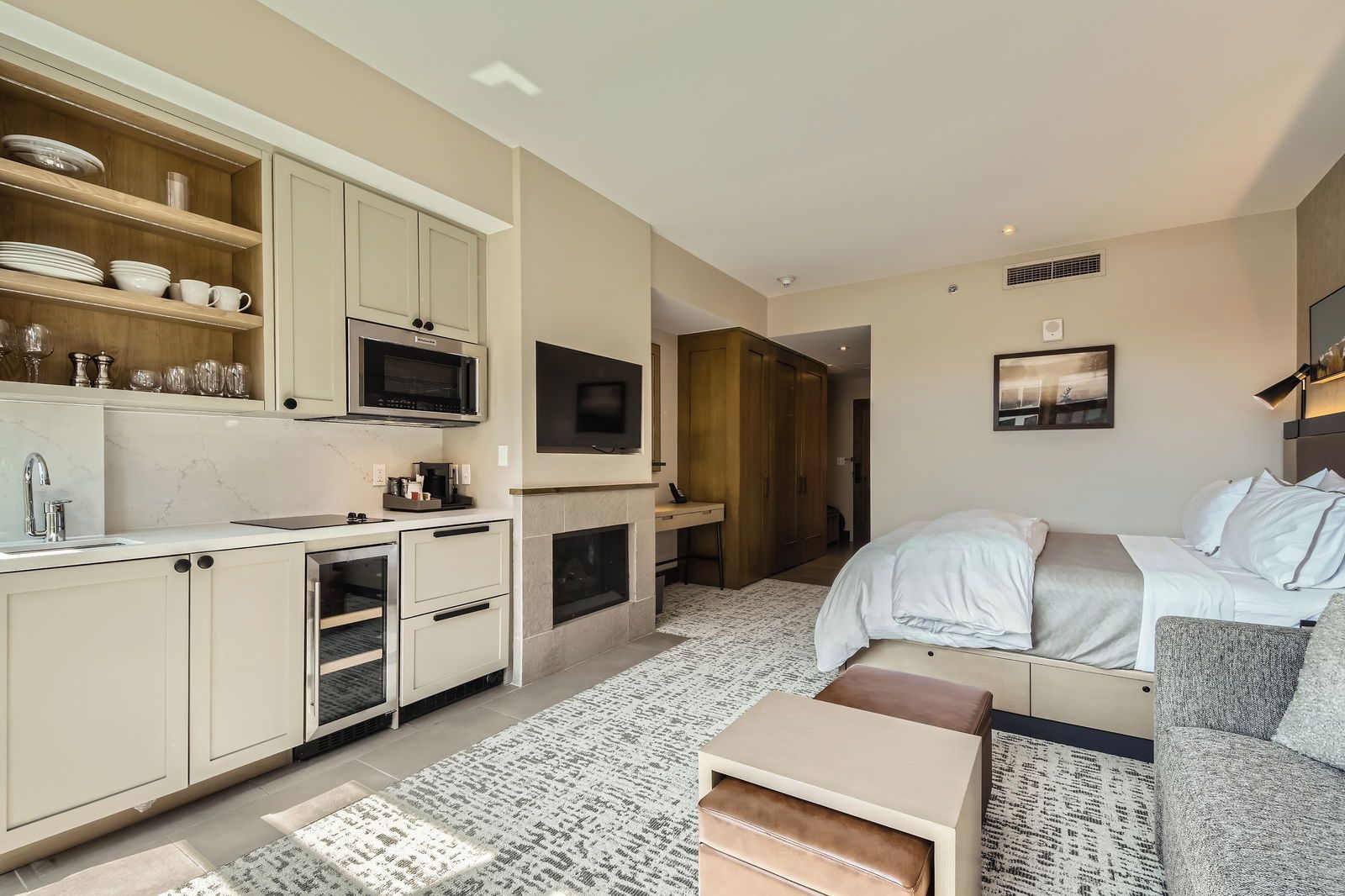 A modern hotel room with a kitchenette, a bed, a sofa, and a fireplace.
