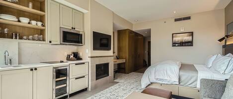 A modern hotel room with a kitchenette, a bed, a sofa, and a fireplace.