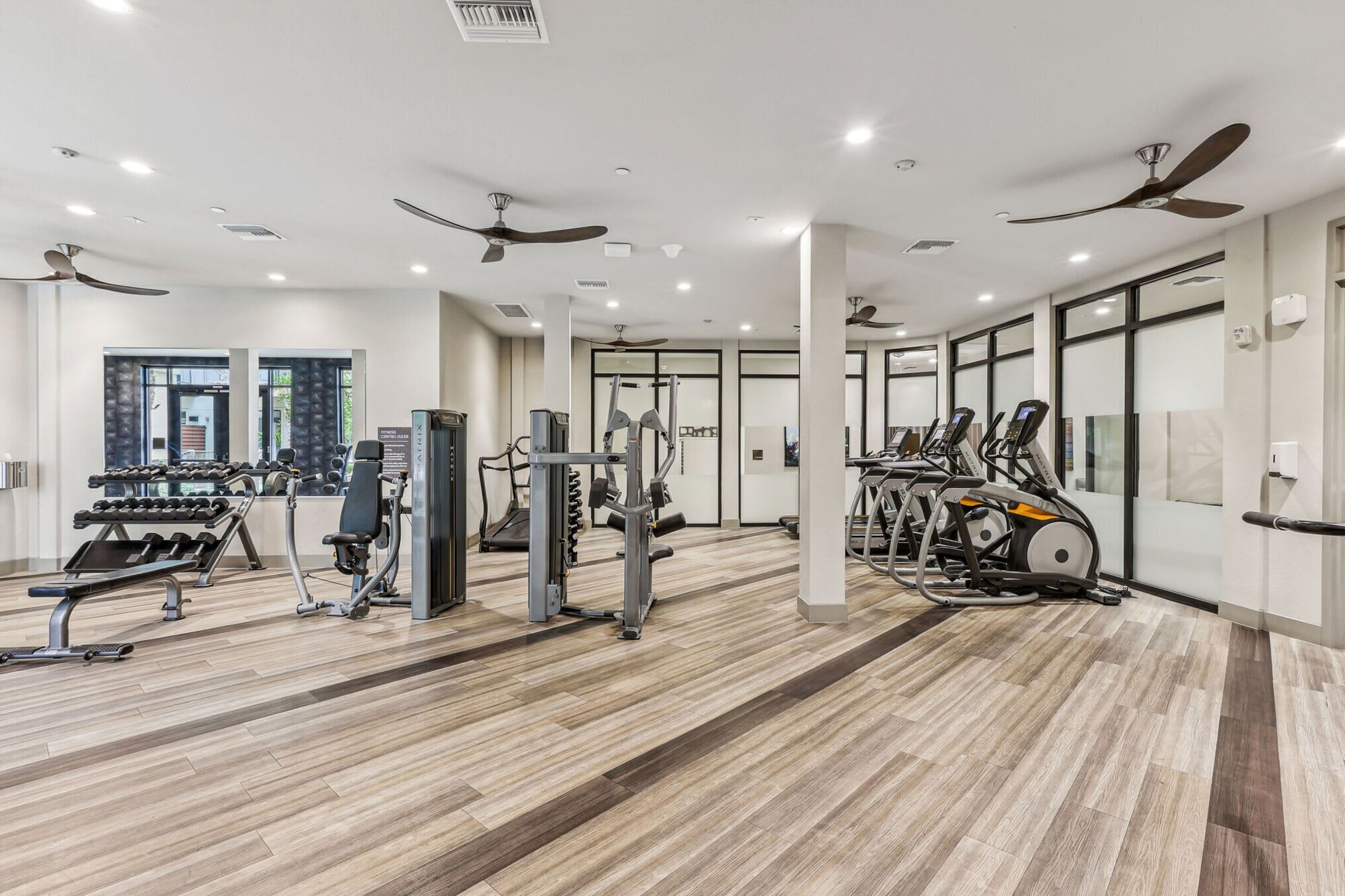 A modern gym with ceiling fans, various exercise equipment, and a wooden floor.