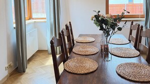 Zona de menjador amb taula de fusta coberta amb sotsplats i cadires, un vas amb flors, i vista d'un edifici a través d'una finestra.