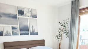 Un dormitori amb un llit gran, terres de fusta, una tauleta de nit, una obra penjada a la paret i una finestra amb cortines.