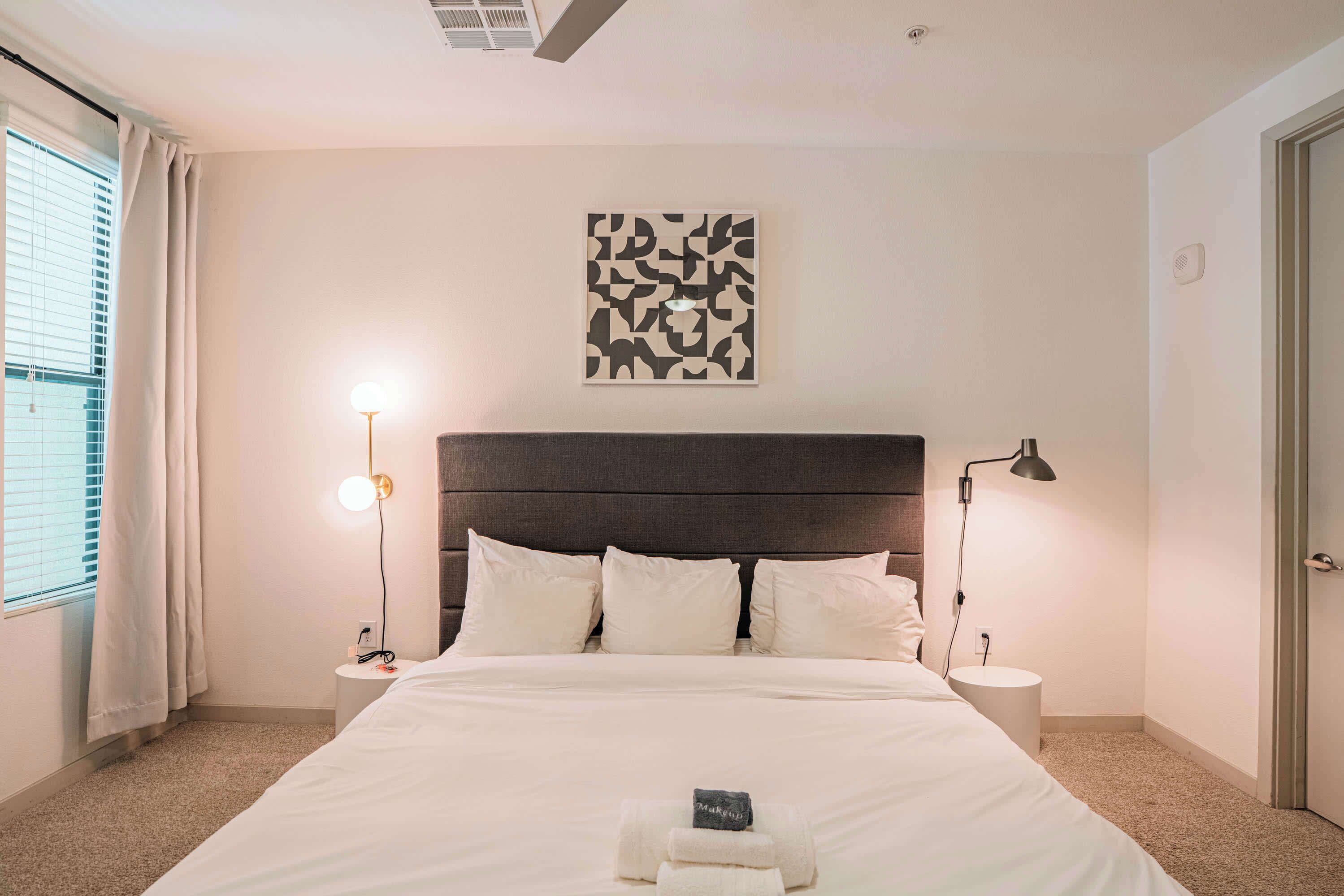A hotel room with a large bed, a headboard, two bedside tables, a wall-mounted artwork, and a window with curtains.