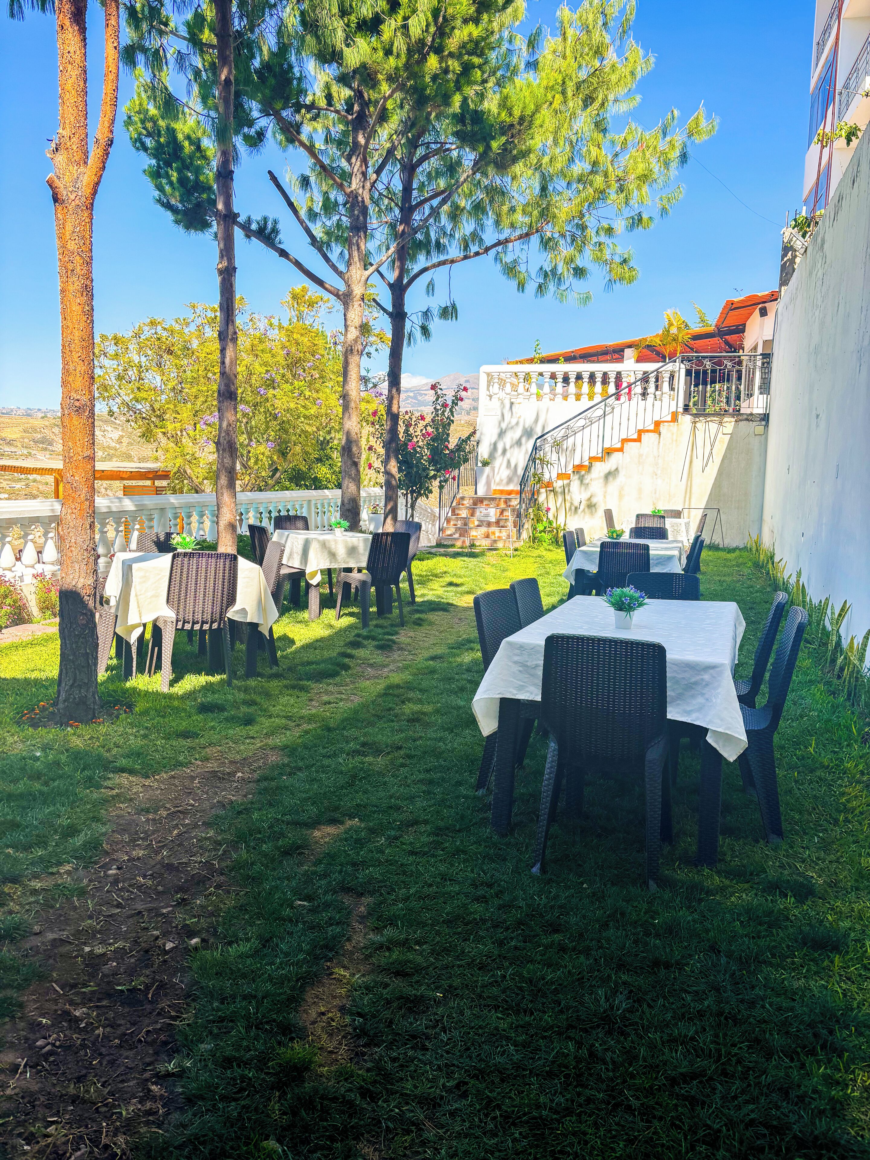 Un espace extérieur aménagé pour les repas, avec des tables et des chaises, entouré d’arbres et offrant une vue sur un bâtiment dont le toit est rouge.