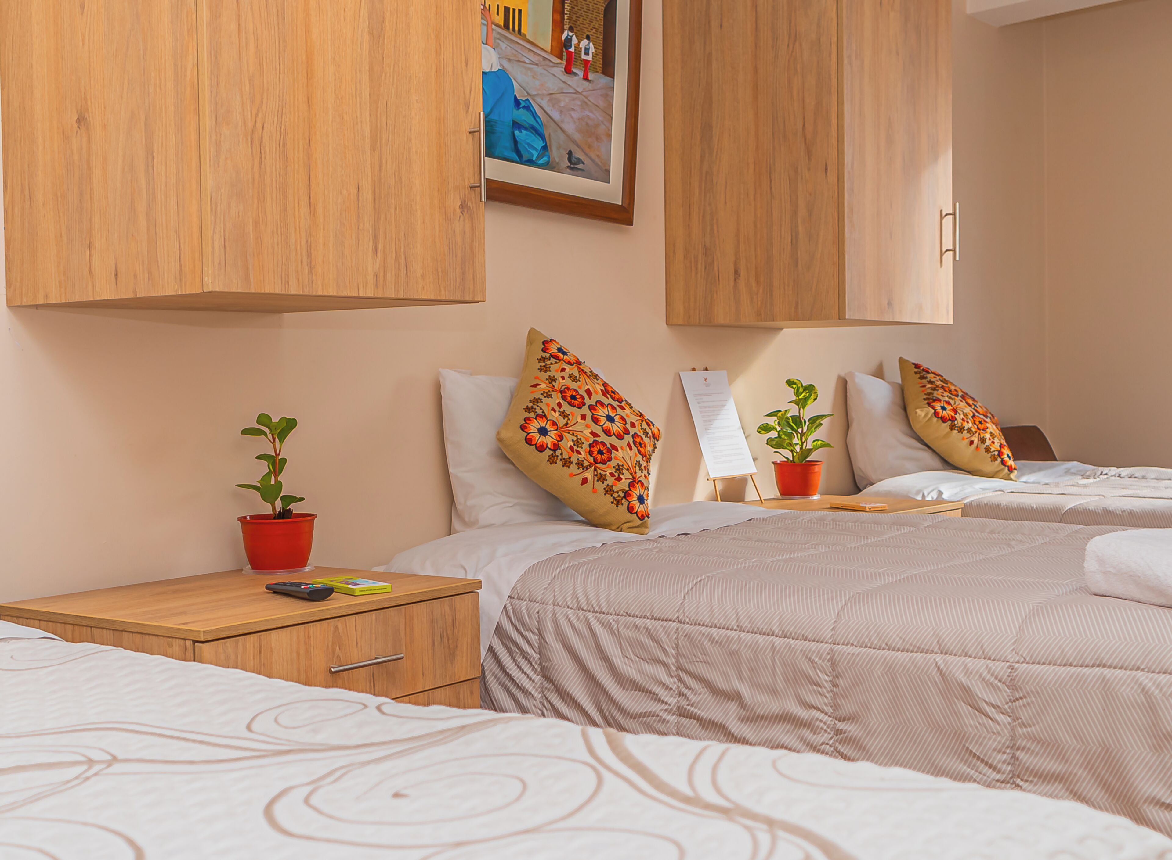 Habitación de hotel con dos camas, armarios de madera, una mesita con una olla roja y un cuadro colgado en la pared.