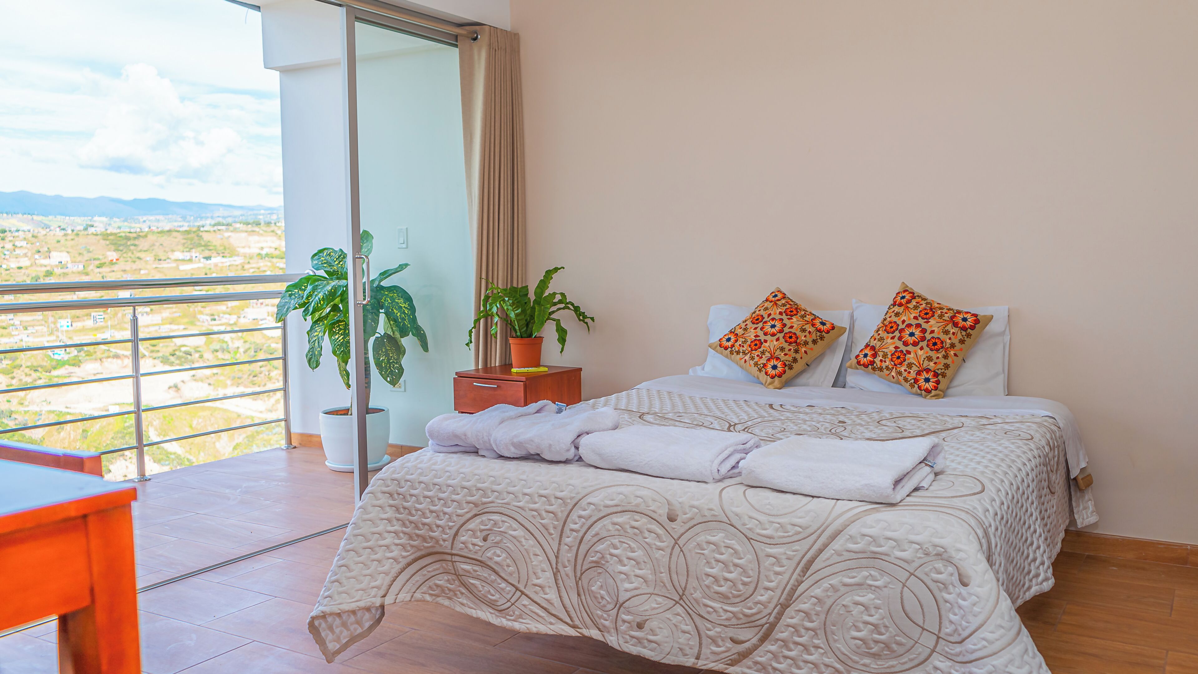 A bedroom with a bed, two pillows, a bedside table with a plant, and a view of the city.