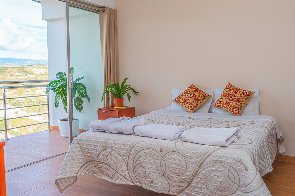 Un dormitorio con cama, dos almohadas, una mesita de noche con una planta y vistas a la ciudad.