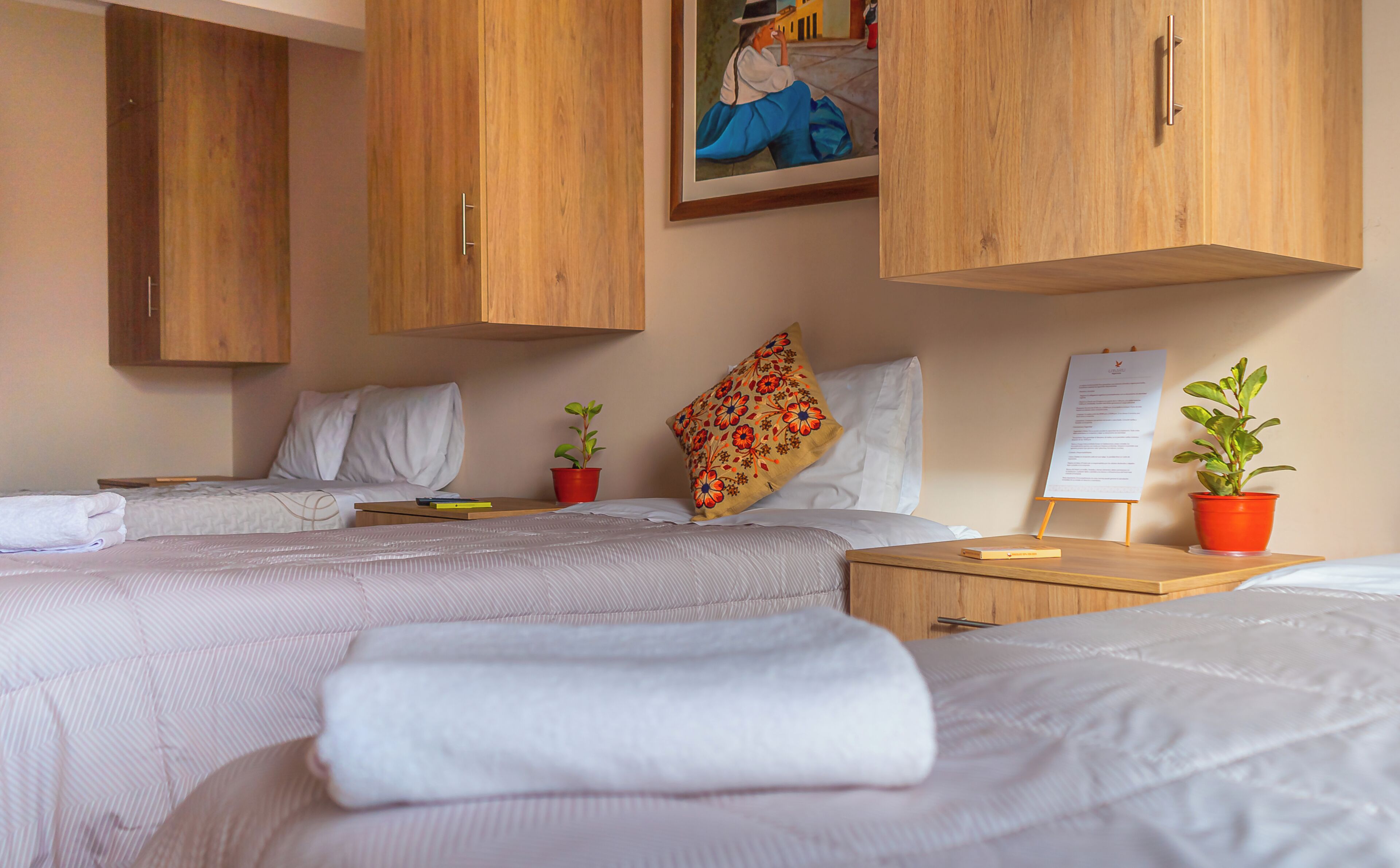 A hotel room with two beds, wooden cabinets, a painting, a bulletin board, and potted plants.