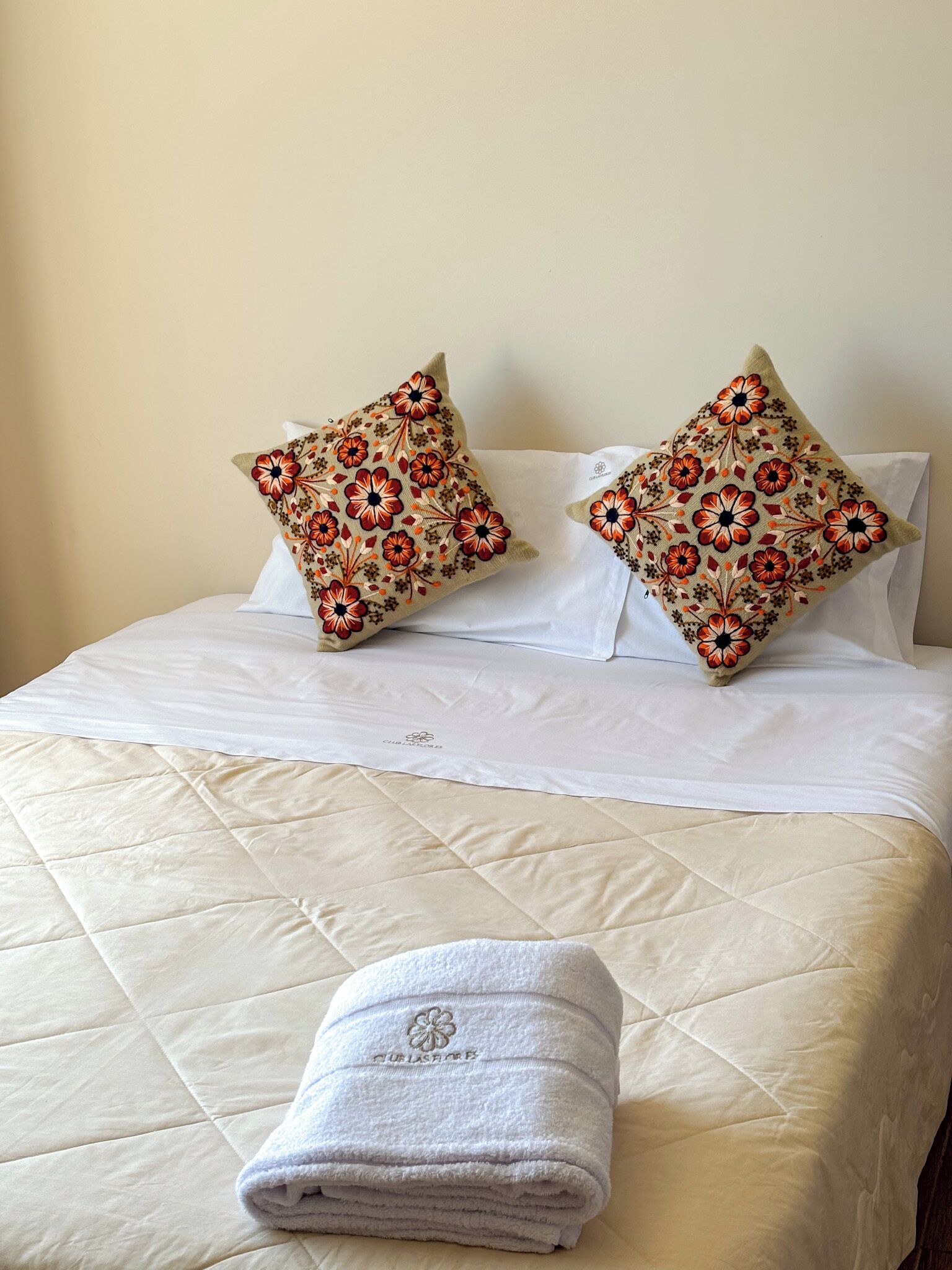 A neatly made bed with a quilted white duvet, two decorative pillows with a floral pattern, and a folded white towel with a logo.