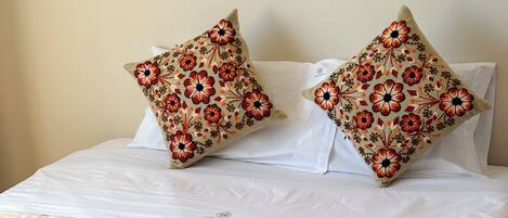 A neatly made bed with a quilted white duvet, two decorative pillows with a floral pattern, and a folded white towel with a logo.