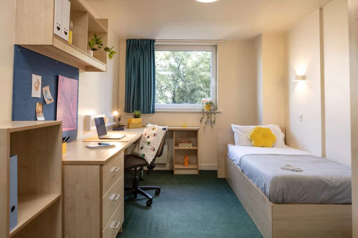 A compact studio apartment with a desk, a bed, a chair, and a small window with curtains.