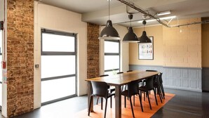 A conference room with a long table, black chairs, pendant lighting, a brick wall, and a large window. - McMillan Student Village in London (London)