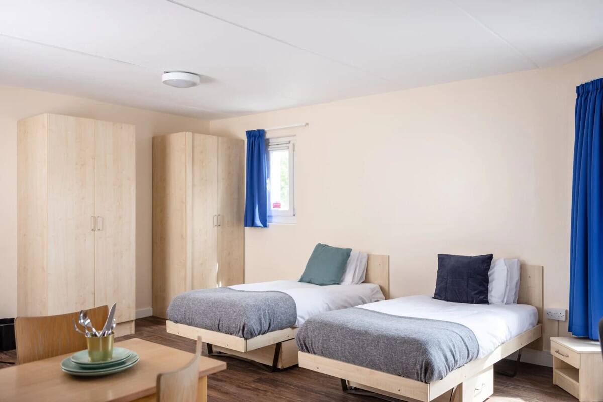 A bedroom with two beds, a wooden wardrobe, a small table with a cup and utensils, and a window with blue curtains.