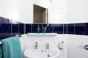 A bathroom with white tiles, a sink, a mirror, a shelf with toiletries, and a towel rack. - McMillan Student Village in London (London)