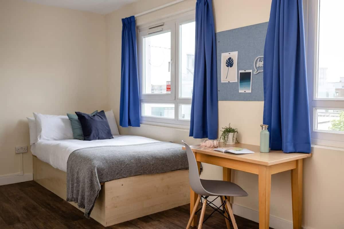 A bedroom with a bed, a desk, and a chair. There are blue curtains on the windows.