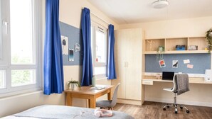 A modern bedroom with a bed, a desk, and a chair. There is a window with curtains, a plant, and a bulletin board with notes. - McMillan Student Village in London (London)