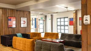 A modern lounge area with wooden panel walls, multiple seating options including orange and gray couches, a red table, and framed artwork on the walls. - McMillan Student Village in London (London)