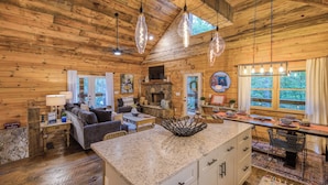 A rustic cabin interior with a stone fireplace, wooden walls, a dining area, and a kitchen with granite countertops. - Whimsical Ways - Hot tub-screen porch-pet friendly (Blue Ridge)