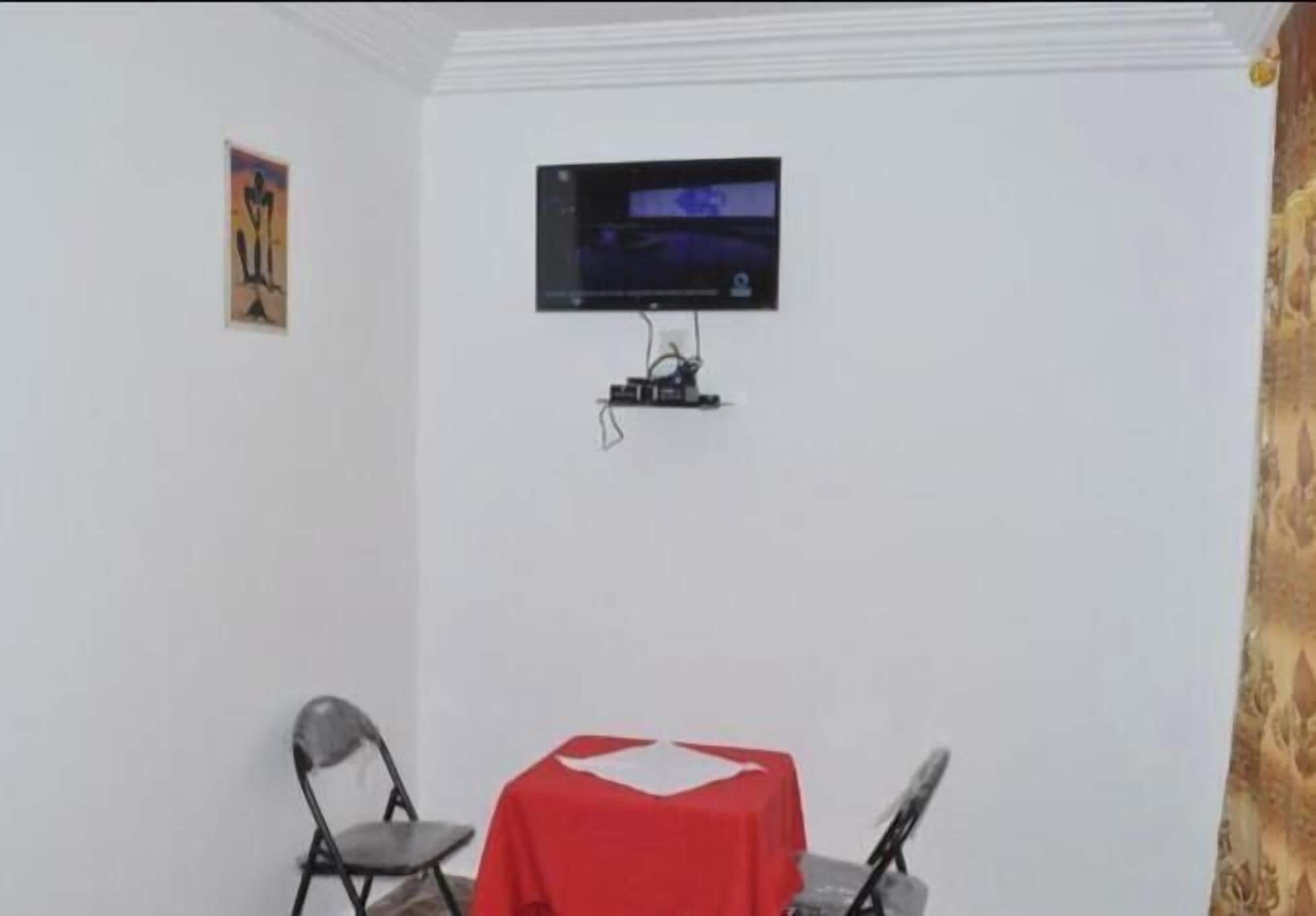 Une petite pièce avec un téléviseur fixé au mur, deux chaises pliantes, une table rouge et un tableau accroché au mur.
