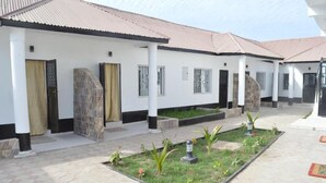 A building with a white exterior, multiple doors, and windows with metal bars. There are small gardens with plants and a paved walkway. - Sukuta Nema Guest House (Sukuta)
