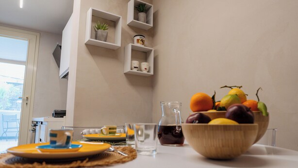 A dining table with a fruit bowl, plates, and cutlery. There are shelves with plants and a small potted plant on the wall. - Beachfront apartment in San Giovanni li Cuti suitable for families (Catania)