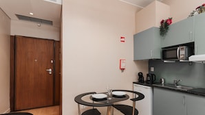 Uma cozinha compacta com micro-ondas, cafeteira e pia. Há uma pequena mesa de jantar com cadeiras e uma porta de madeira.