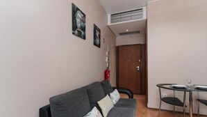 Uma sala de estar moderna com um sofá cinzento, uma pequena mesa de jantar com cadeiras e uma porta de madeira ao fundo.
