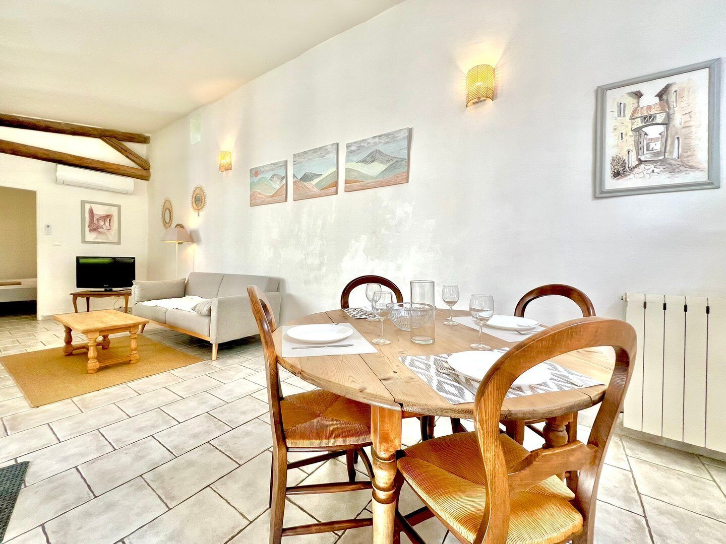 Zona pranzo con tavolo rotondo in legno apparecchiato per quattro, circondato da sedie in legno con sedili in vimini. Tre opere d'arte incorniciate sono appese alla parete sopra il tavolo, e si nota un termosifone a destra.