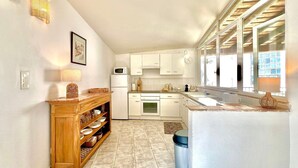 Una cocina con gabinetes blancos, un microondas y un lavamanos. Se observa una estantería de madera con vajilla y un bote de basura.