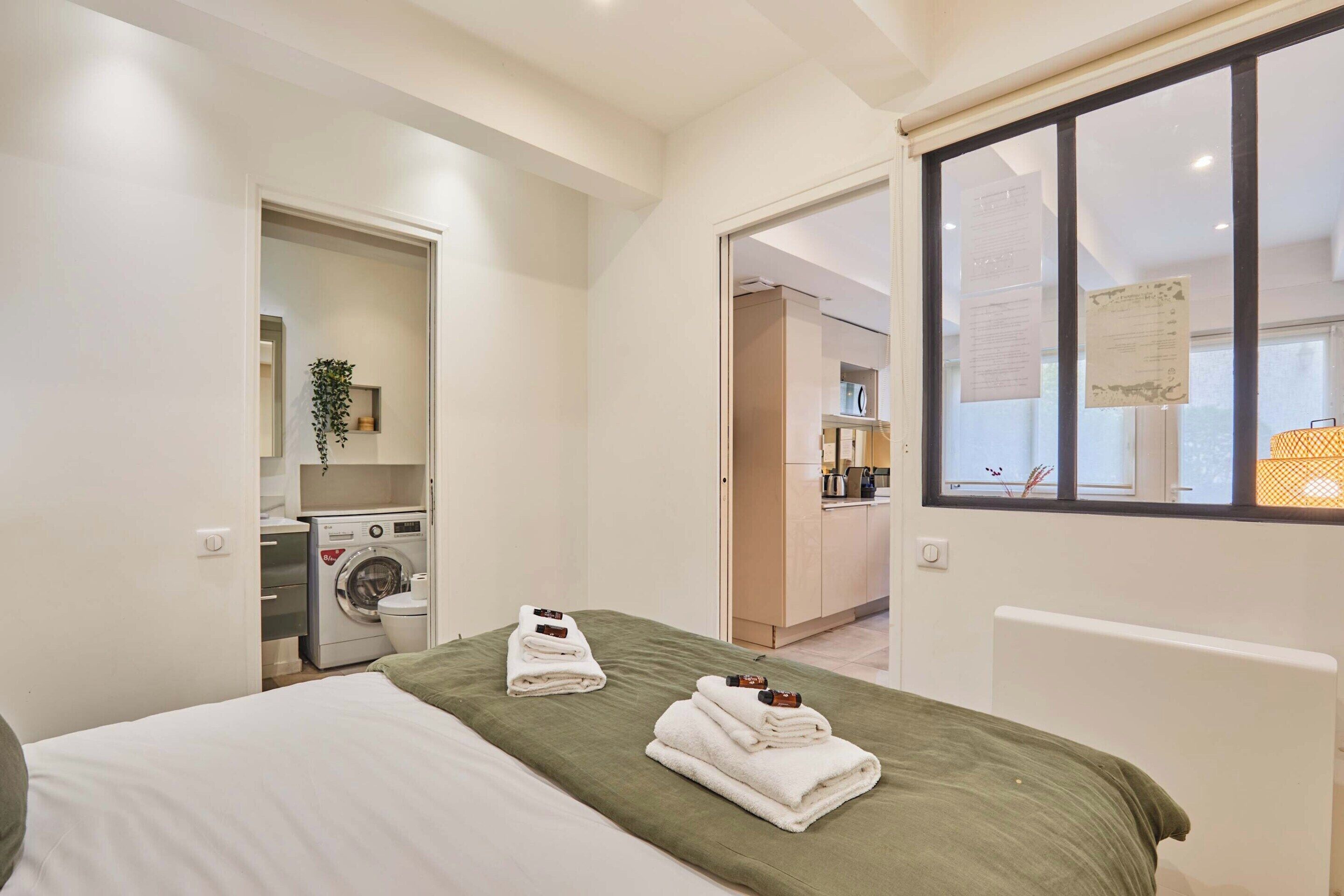 A hotel room with a bed, a green bedspread, neatly folded towels, a washing machine, and a view of the kitchen through an open door.