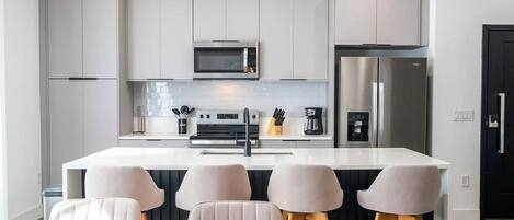 A modern kitchen with white cabinetry, a stainless steel refrigerator, a microwave, and a coffee machine. There are four bar stools and a white dining table.