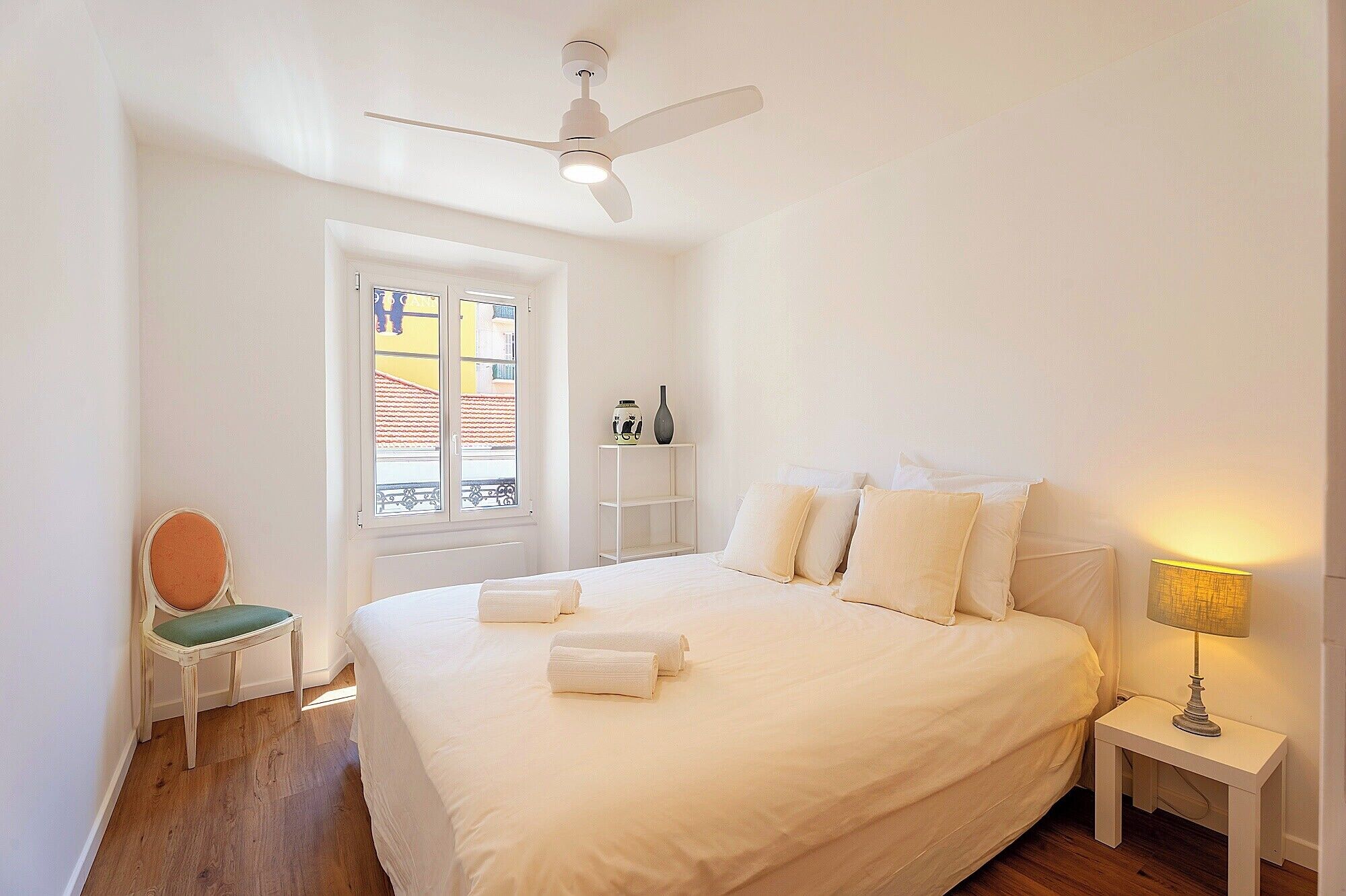 Ein Schlafzimmer mit Bett, Stuhl, Nachttisch, Lampe, Deckenventilator und Fenster mit Blick auf Gebäude.