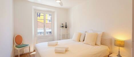A bedroom with a bed, a chair, a nightstand, a lamp, a ceiling fan, and a window with a view of buildings.