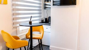 A small dining area with a round table and two yellow chairs, a kitchen area with a stove and cabinets, and a large window with blinds.