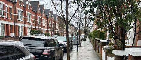 A residential street with parked cars, a sidewalk, and houses with red brick facades.